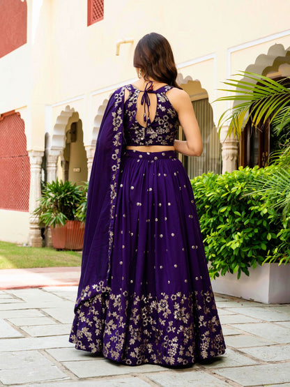 Back view of model wearing royal purple lehenga with cut-out tie-up blouse and floral-bordered pleated skirt