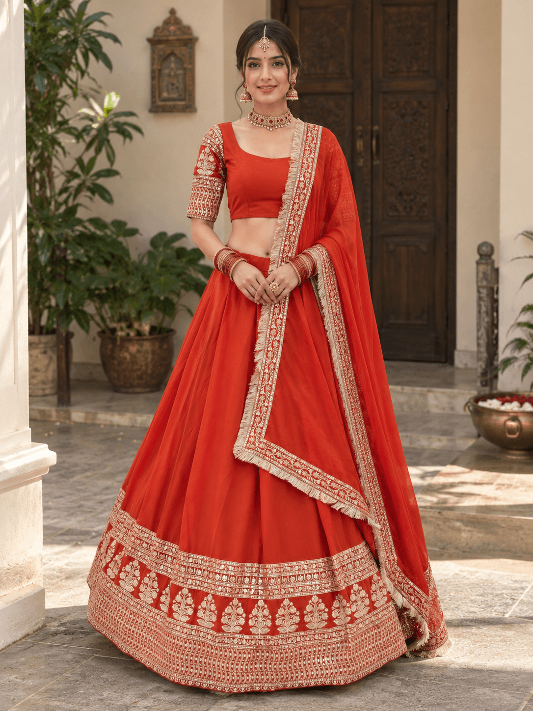 Full front view of model in a rust orange flared lehenga choli with gold tree motif border and fringe dupatta