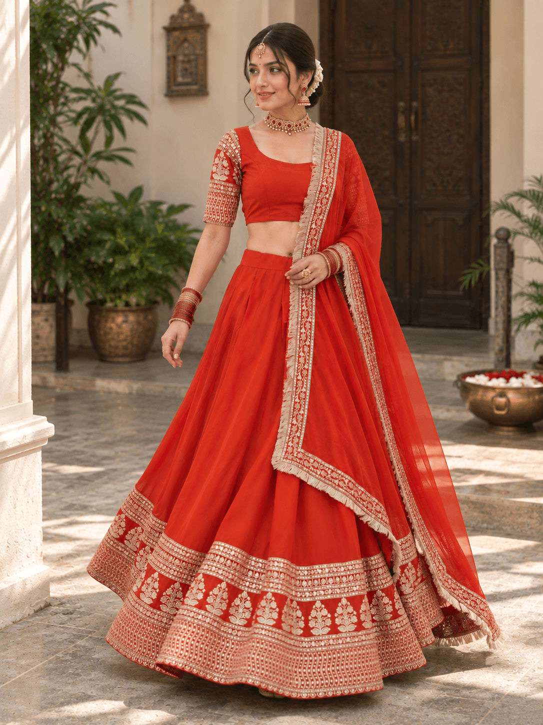 Model standing in a rust red pleated lehenga featuring wide gold decorative hemline embroidery