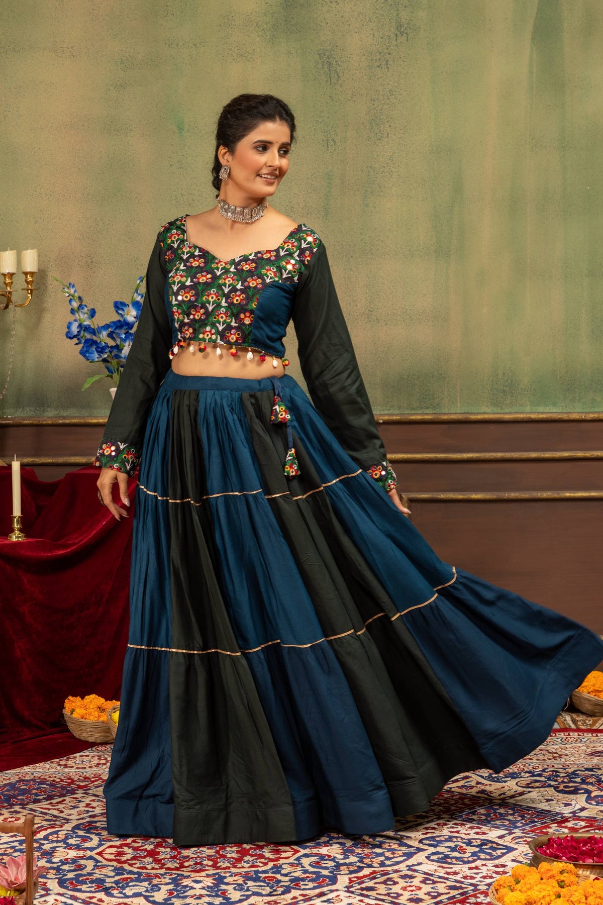 Three-quarter standing pose of model in tiered teal and dark green lehenga skirt with embroidered full-sleeve choli