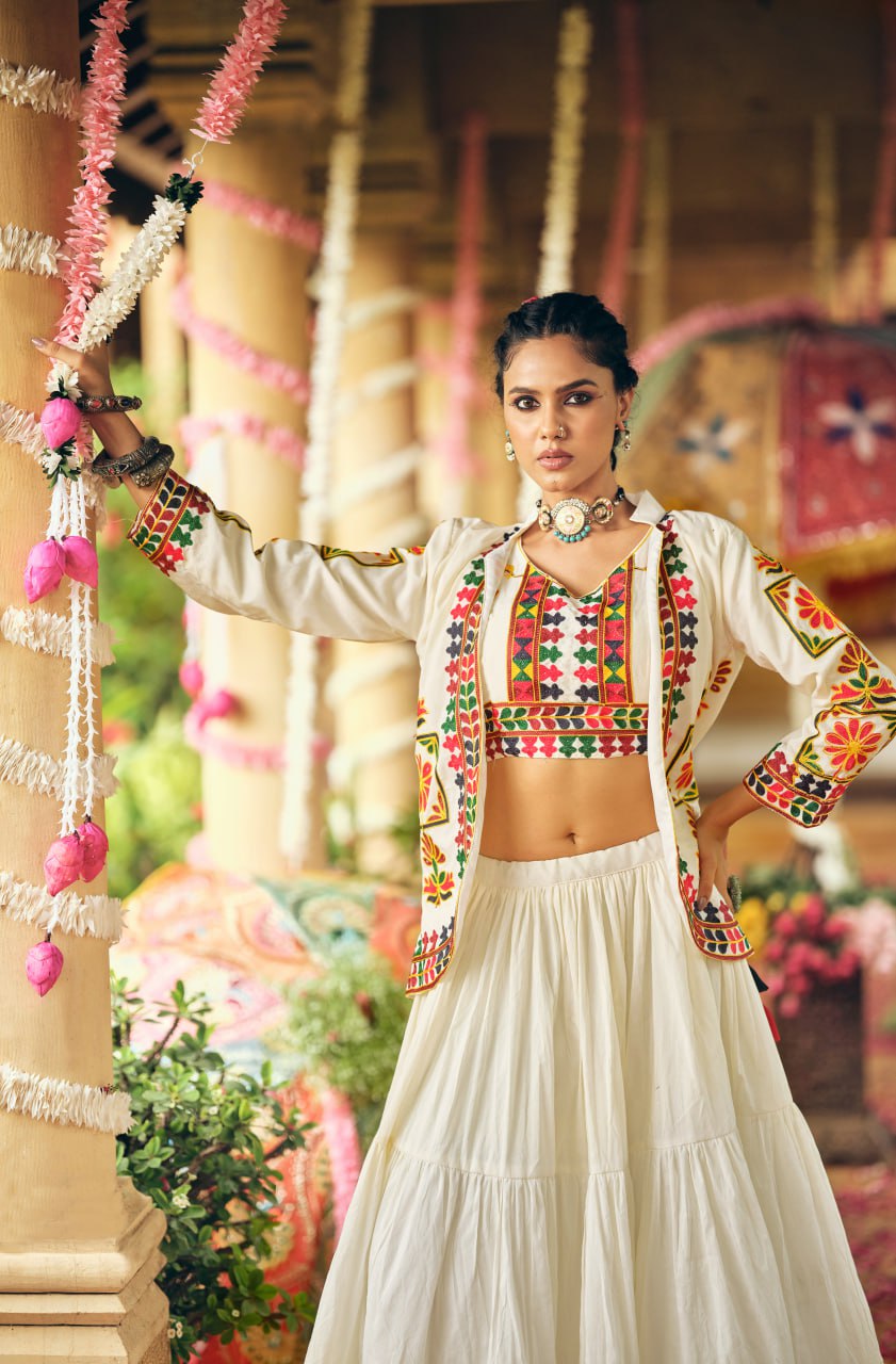 Model leaning against a temple pillar in a white embroidered crop top, matching embroidered jacket, and tiered flared skirt