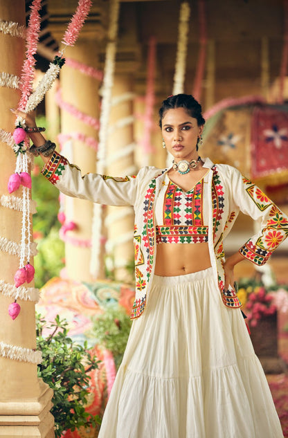Model leaning against a temple pillar in a white embroidered crop top, matching embroidered jacket, and tiered flared skirt