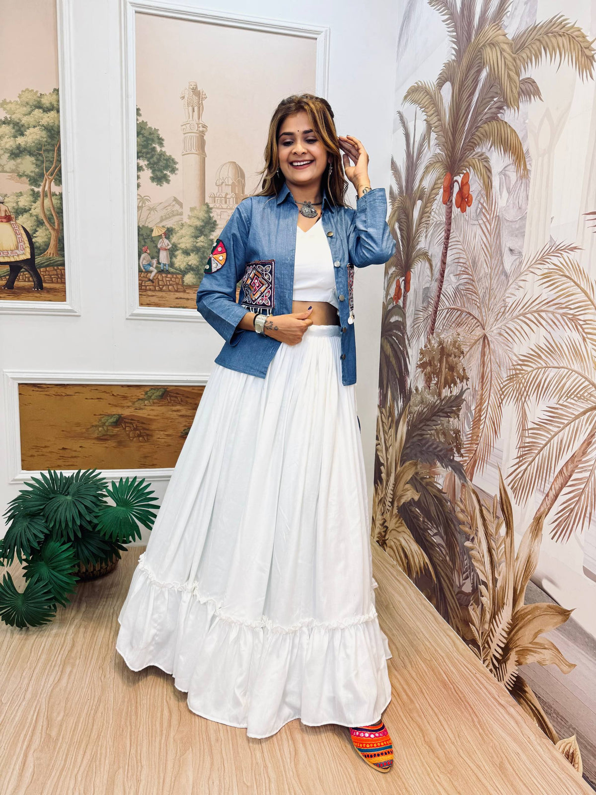 Standing full-length front view of a model in a white tiered ruffle lehenga and blue jacket with front pocket embroidery