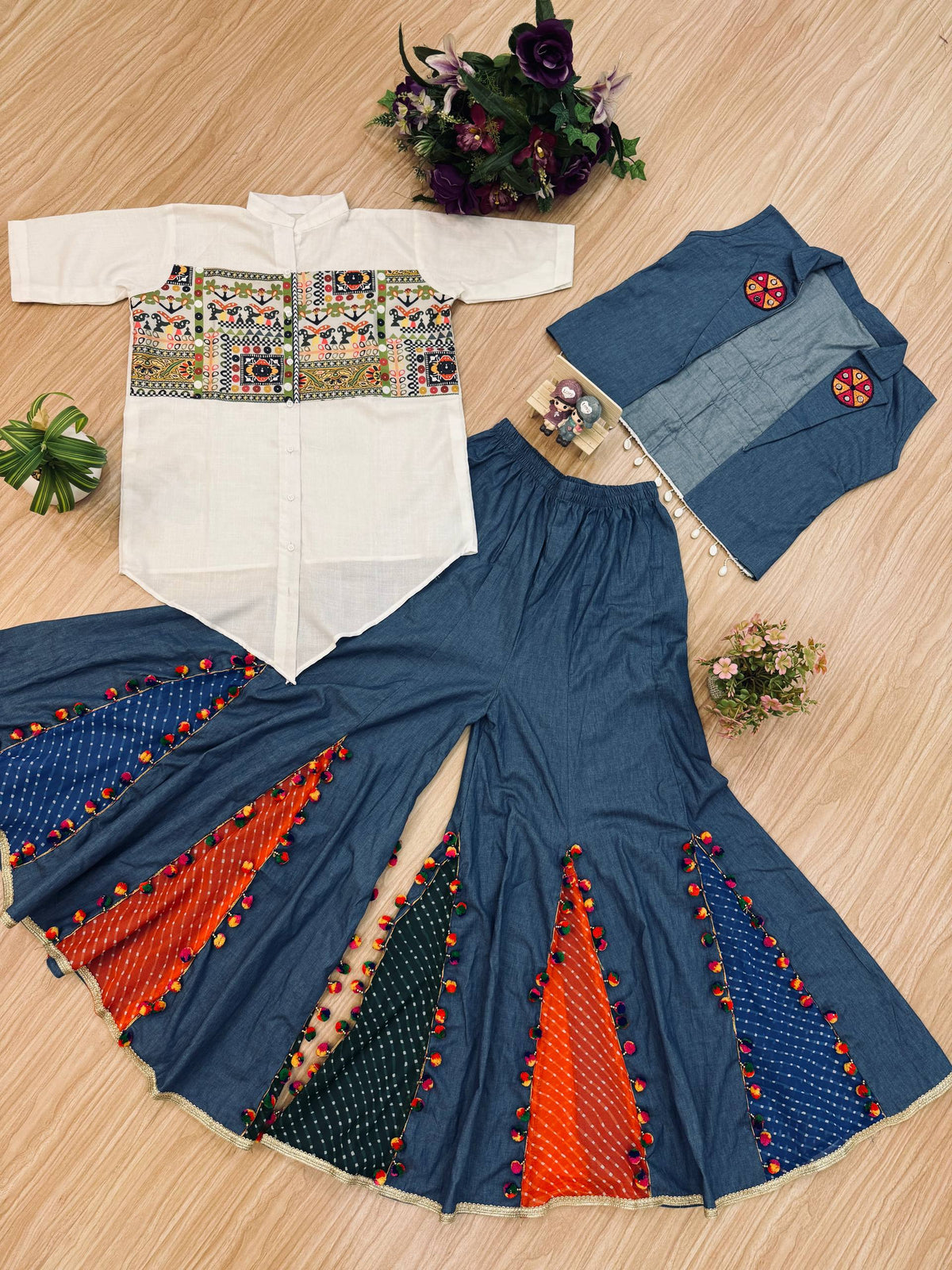 Flat lay view of white embroidered kediya shirt, blue sleeveless jacket, and blue flared sharara pants with colorful inserts