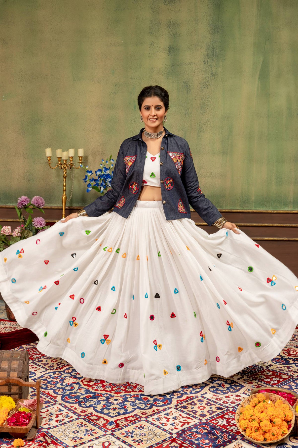 Model holding out flared white lehenga skirt with colorful mirror accents paired with embroidered blue jacket