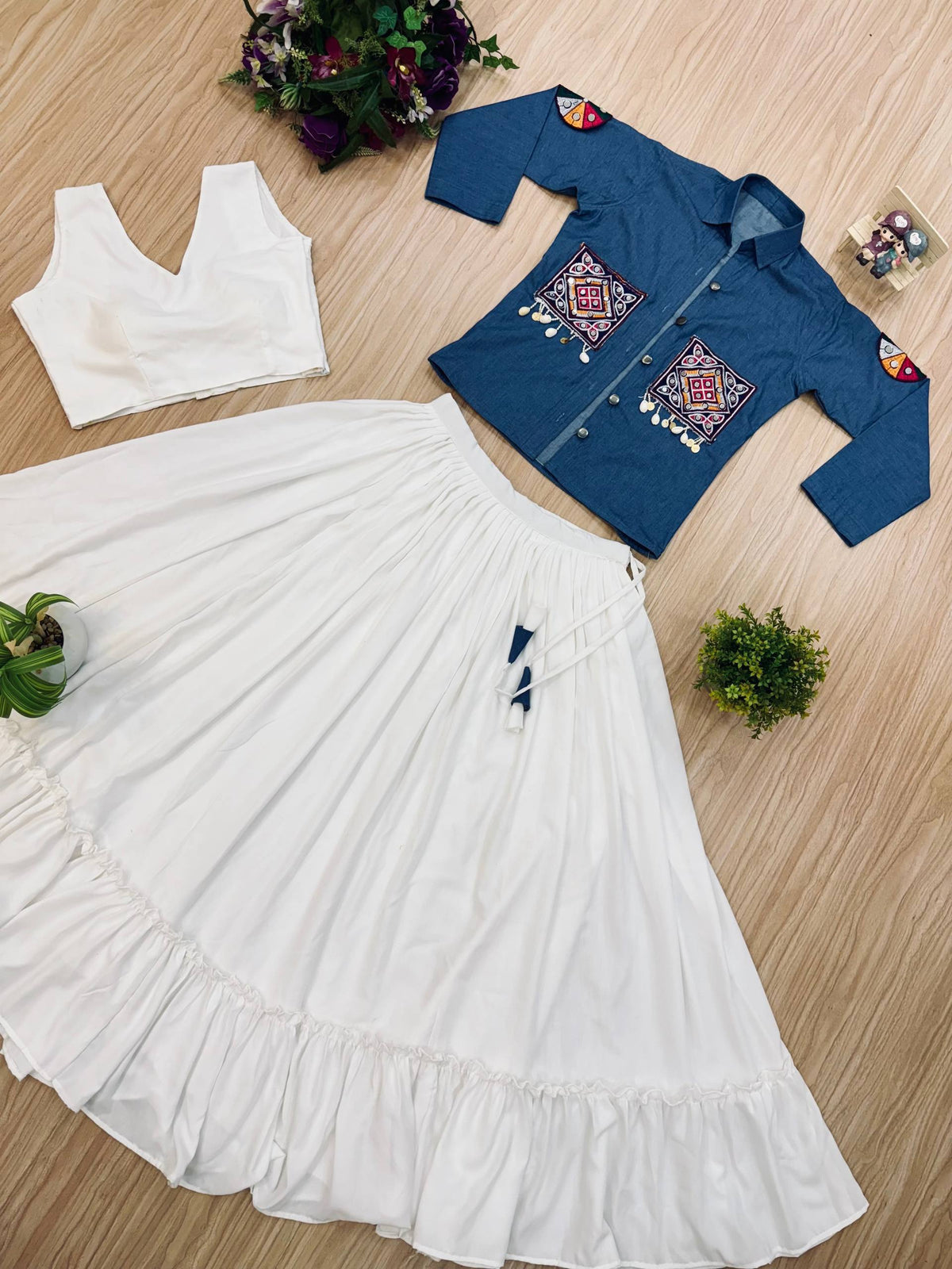 Flat lay of a white flared ruffle lehenga skirt paired with a sleeveless crop top and blue embroidered jacket with coin drops