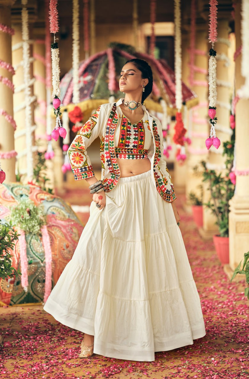 Full-length front view of a model in a tiered white lehenga skirt, embroidered crop top, and full-sleeve white jacket with tassels